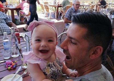 Smiling baby wearing a pink headband held by an adult at an outdoor cafe
