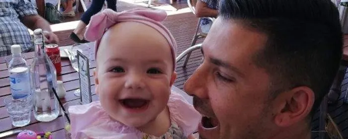 Smiling baby wearing a pink headband held by an adult at an outdoor cafe