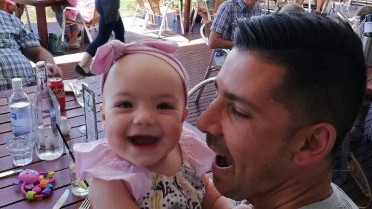 Smiling baby wearing a pink headband held by an adult at an outdoor cafe