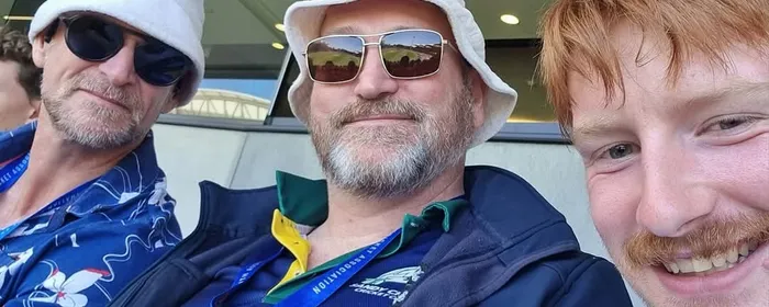 Three people in stadium seating, wearing hats and sunglasses, smiling at the camera.