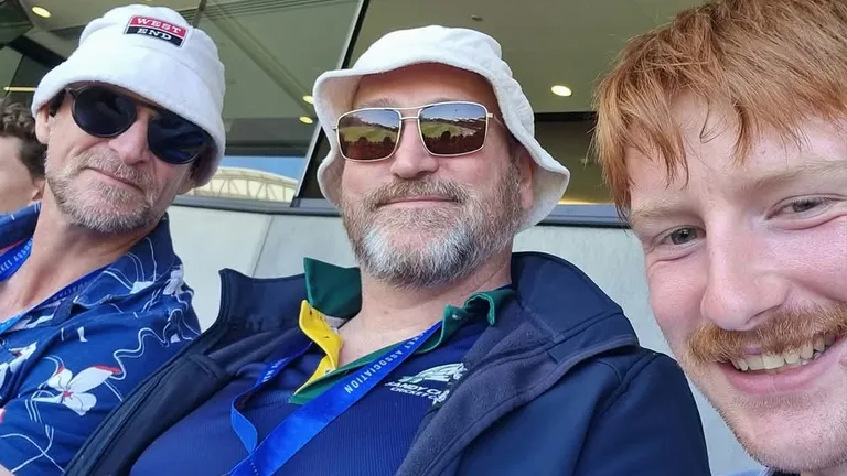 Three people in stadium seating, wearing hats and sunglasses, smiling at the camera.