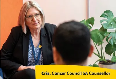 Counsellor with blonde hair and glasses in a black blazer speaks to a client; Cancer Council SA sign.