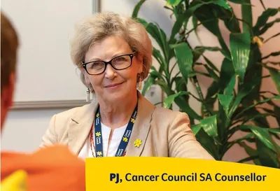 A person with short light hair and glasses sits at a table with a yellow Cancer Council SA sign.