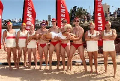 Group of people in swimsuits on a beach posing with a white dog, red banners in the background.