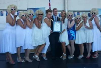 Group in white dresses posing on stage as a person in a suit holds a framed certificate.