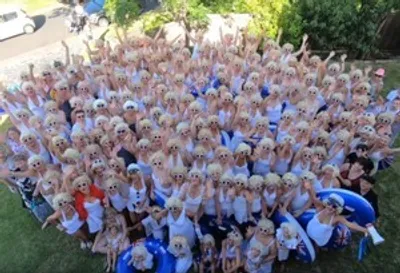 Large group of people outdoors in light clothing, many with blonde wigs, posing on a lawn.