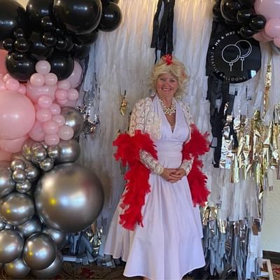 Person in lavender dress with red feather boa stands before a silver fringe backdrop with balloons.
