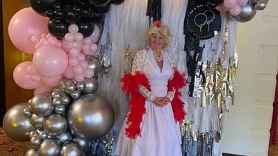 Person in lavender dress with red feather boa stands before a silver fringe backdrop with balloons.