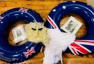 Two blue Australian-flag lifebuoys on a wooden floor, a blonde wig with sunglasses, a white robe, and framed photos.