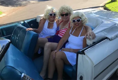 Three people in white outfits and blonde wigs riding in a blue vintage convertible.