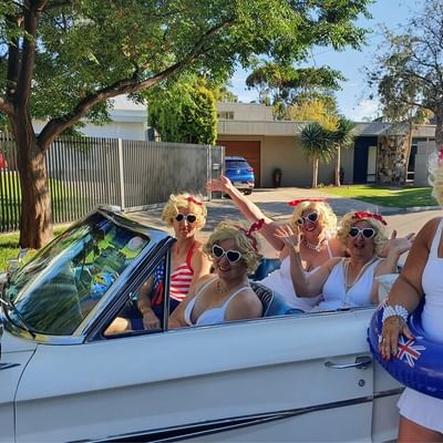Group in white retro outfits with blonde wigs and heart sunglasses ride in a vintage convertible.