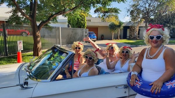 Group in white retro outfits with blonde wigs and heart sunglasses ride in a vintage convertible.