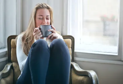 Person with long blonde hair sits in a vintage armchair by a window, sipping from a mug.