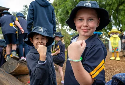 Two kids in navy uniforms and hats pose with fists at a playground; yellow mascot in background.