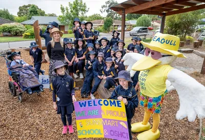 Students in navy uniforms and hats pose with a yellow pelican mascot at a school playground.