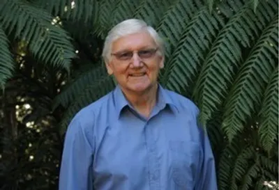 Smiling older person with white hair and glasses in a light blue shirt, in front of fern leaves.