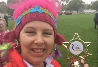 Smiling person wearing a pink knitted hat with blue flowers, holding a star trophy on a grassy field.
