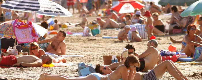 Bustling beach scene with many sunbathers on the sand beneath colourful umbrellas.