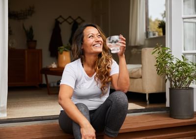 Person sitting on a wooden doorstep at an open doorway, smiling, holding a glass of water.