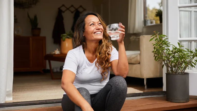 Person sitting on a wooden doorstep at an open doorway, smiling, holding a glass of water.