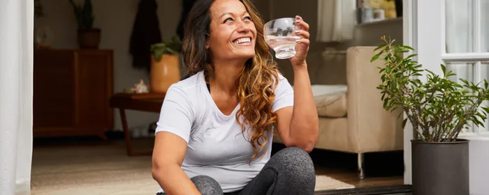 Person sitting on a wooden doorstep at an open doorway, smiling, holding a glass of water.