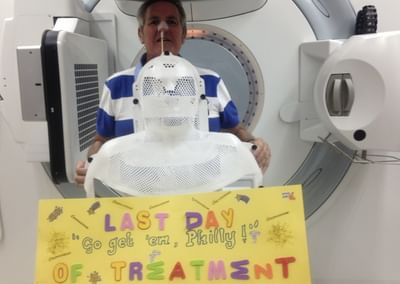 A person wearing a white mesh mask stands in front of an MRI machine; a yellow sign reads Last Day of Treatment.