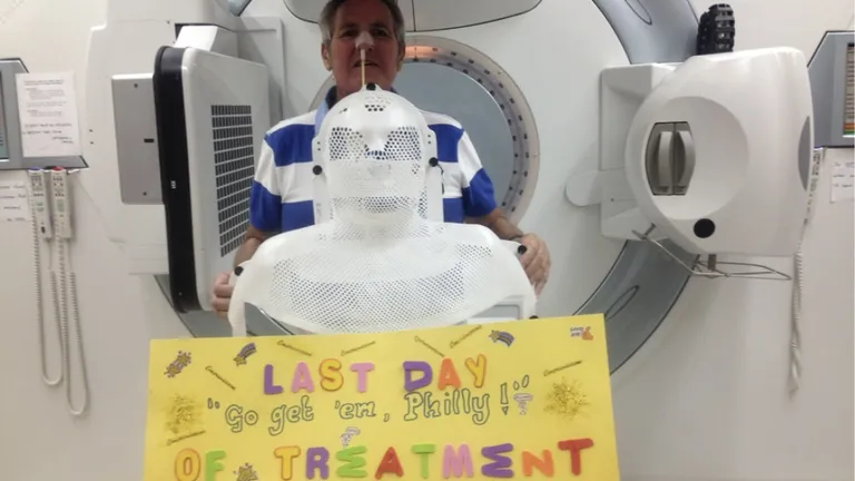 A person wearing a white mesh mask stands in front of an MRI machine; a yellow sign reads Last Day of Treatment.