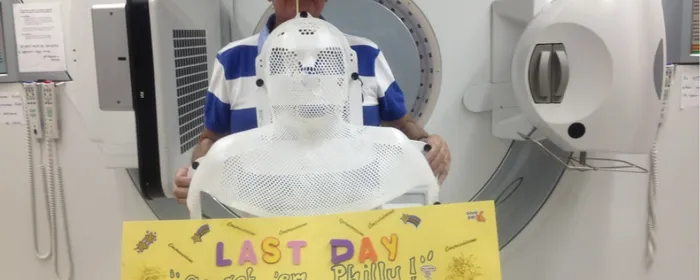 A person wearing a white mesh mask stands in front of an MRI machine; a yellow sign reads Last Day of Treatment.