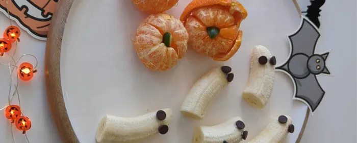 Halloween-themed plate with banana ghosts, pumpkin-shaped mandarins, and a black spider decoration.
