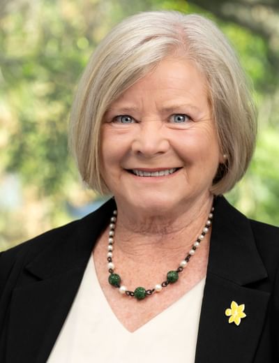Older person with grey hair in a black blazer, green beaded necklace and flower pin, smiling.