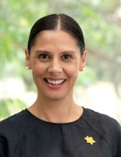 Smiling person with dark hair tied back, wearing a black top with a yellow pin, outdoors.