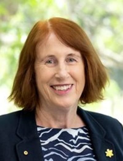 Smiling person with shoulder-length red hair in a dark blazer and patterned blouse, outdoors.