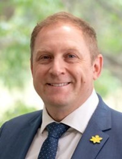 Smiling person in a blue suit and tie with a yellow star lapel pin, outdoors.