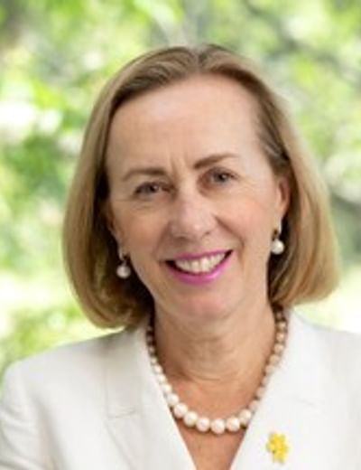 Smiling person in a white blazer with a pearl necklace, outdoors with greenery.