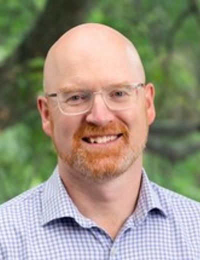 Smiling bald person wearing glasses and a light blue checked shirt, outdoors among greenery.