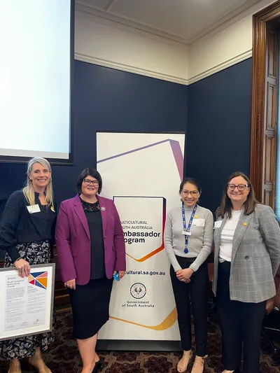 Four people stand indoors beside a Multicultural South Australia Ambassador Program banner, smiling.