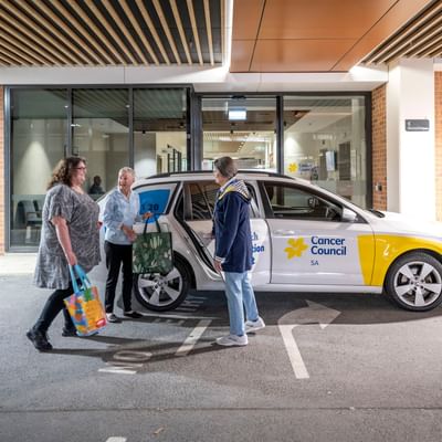 Three people stand beside a white Cancer Council car in a car park outside a glass-front building.
