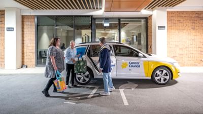 Three people stand beside a white Cancer Council car in a car park outside a glass-front building.
