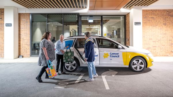 Three people stand beside a white Cancer Council car in a car park outside a glass-front building.