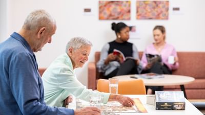 Two older adults working on a jigsaw puzzle at a bright table, with two readers on a sofa in the background.
