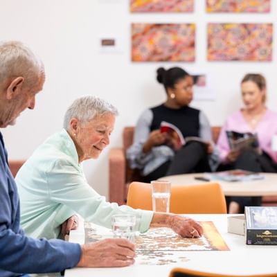 Two older adults working on a jigsaw puzzle at a bright table, with two readers on a sofa in the background.