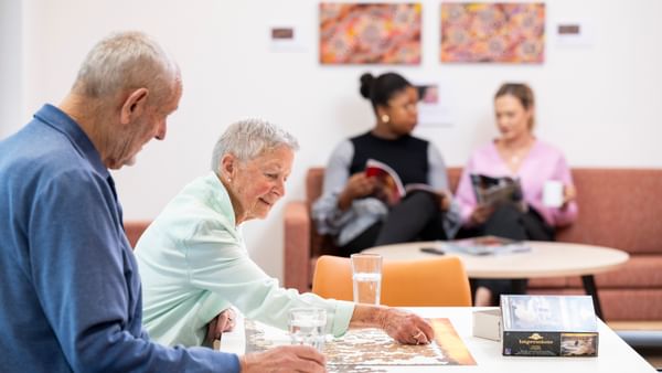 Two older adults working on a jigsaw puzzle at a bright table, with two readers on a sofa in the background.