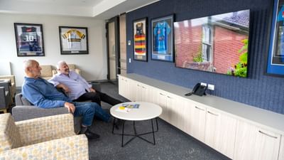 Two people sit on chairs in a lounge with framed sports jerseys on the wall and a round table.