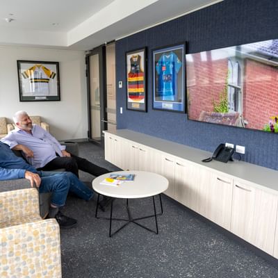 Two people sit on chairs in a lounge with framed sports jerseys on the wall and a round table.