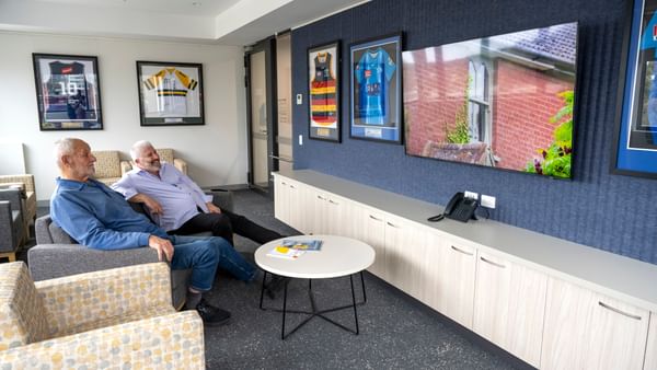 Two people sit on chairs in a lounge with framed sports jerseys on the wall and a round table.