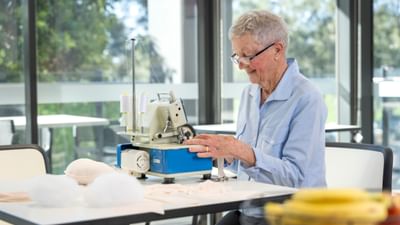 Older person with short grey hair and glasses sewing on a sewing machine at a bright table.