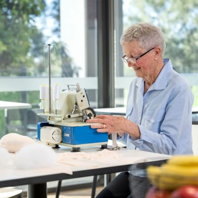 Older person with short grey hair and glasses sewing on a sewing machine at a bright table.