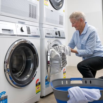 Elderly person loading a front-loading washing machine in a laundromat, with a blue laundry basket.