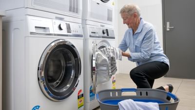Elderly person loading a front-loading washing machine in a laundromat, with a blue laundry basket.
