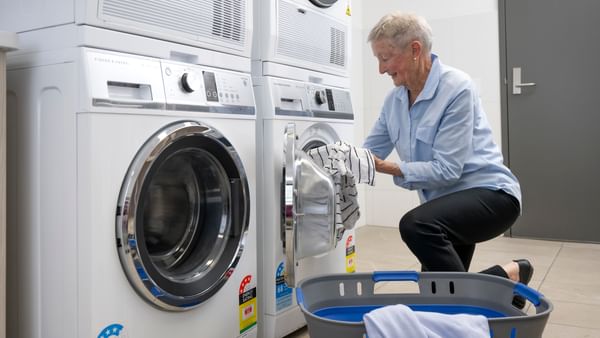 Elderly person loading a front-loading washing machine in a laundromat, with a blue laundry basket.
